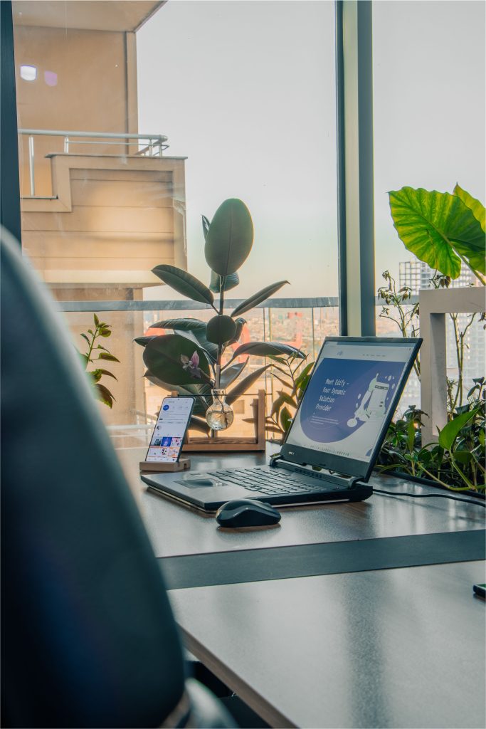 Contemporary office setup featuring a laptop and plants, illuminated by natural light through large windows.